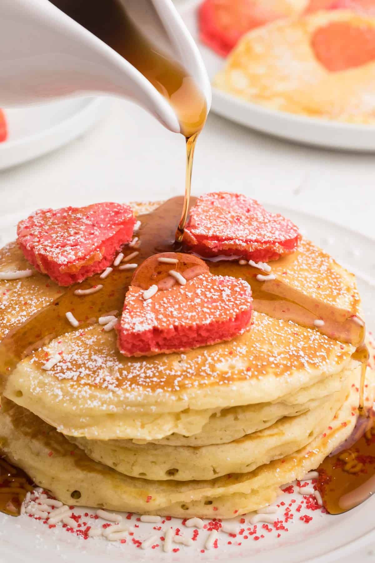 Du sirop est versé sur des pancakes avec d'autres pancakes en forme de cœur au-dessus.