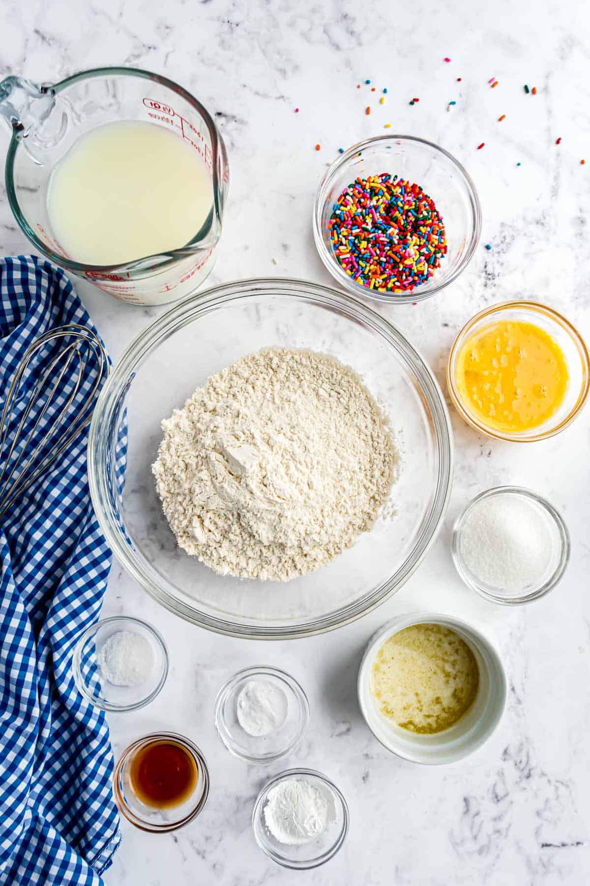 Bols en verre clair avec des ingrédients : farine, œufs, vanille, lait, confettis, et plus.