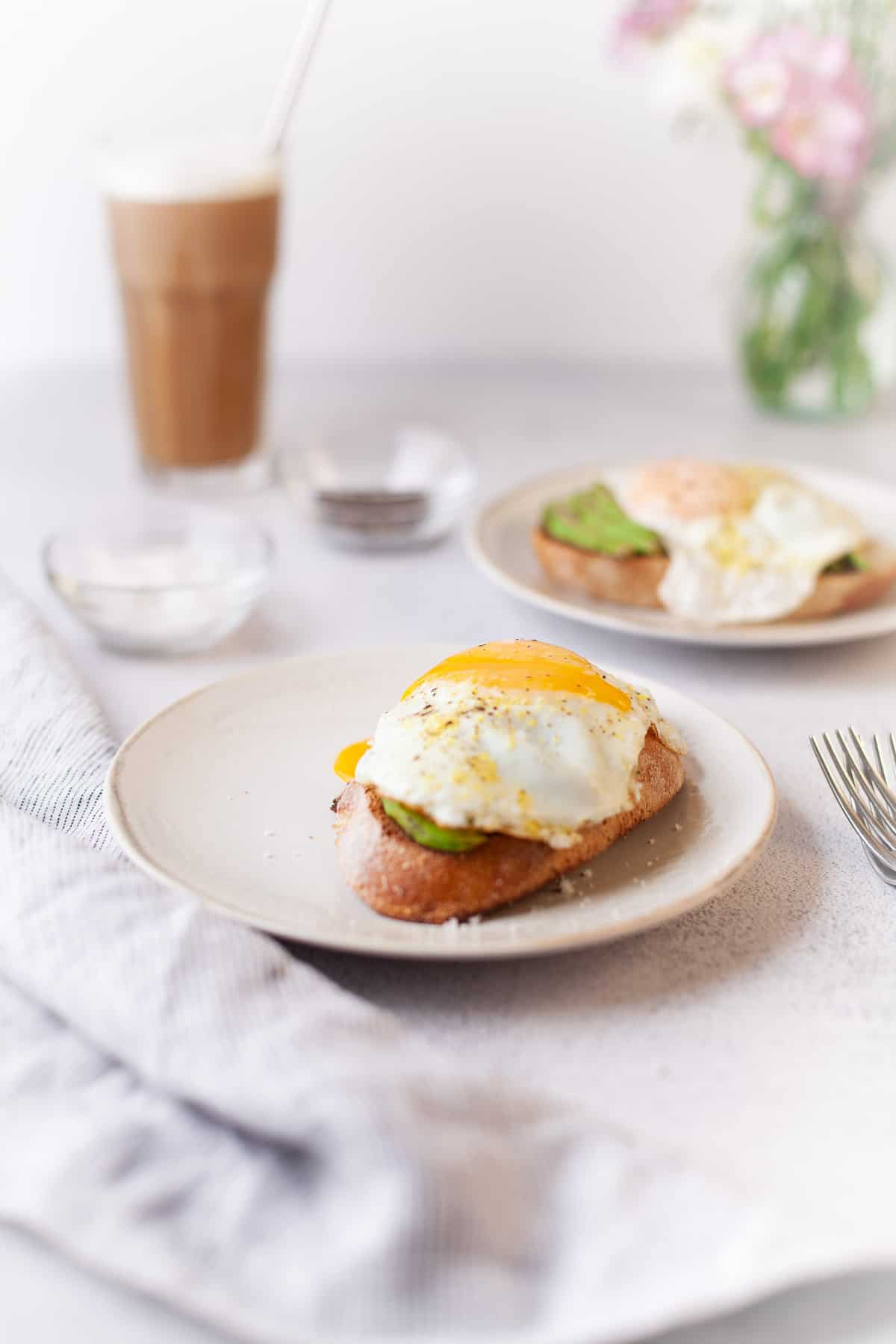 Tableau blanc avec deux toasts à l’avocat garnis d'œufs, du café et un vase de fleurs.