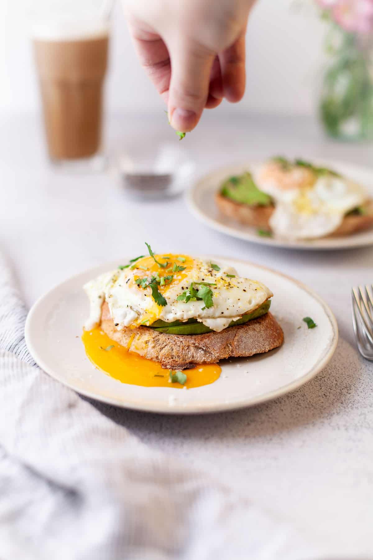 Toast à l’avocat garnie d’un œuf en train d’être saupoudrée d’herbes.