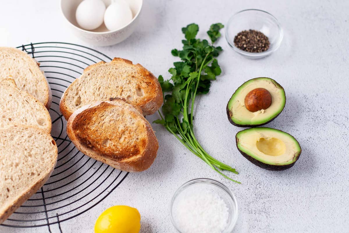 Toast, herbes, avocats, œufs et assaisonnements sur une surface grise.
