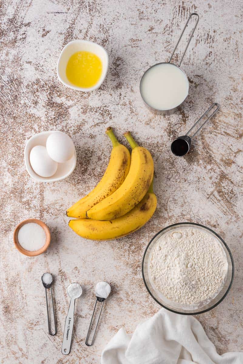 Ingrédients sur une surface beige et blanche : farines, sucres, bananes mûres, œufs, huile, extrait de vanille.