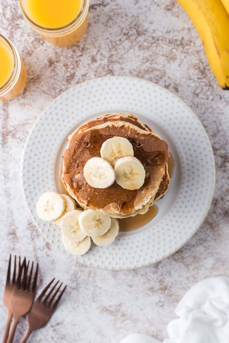 Vue de dessus des pancakes à la banane. Du jus d'orange, des fourchettes, et une banane sont également visibles.