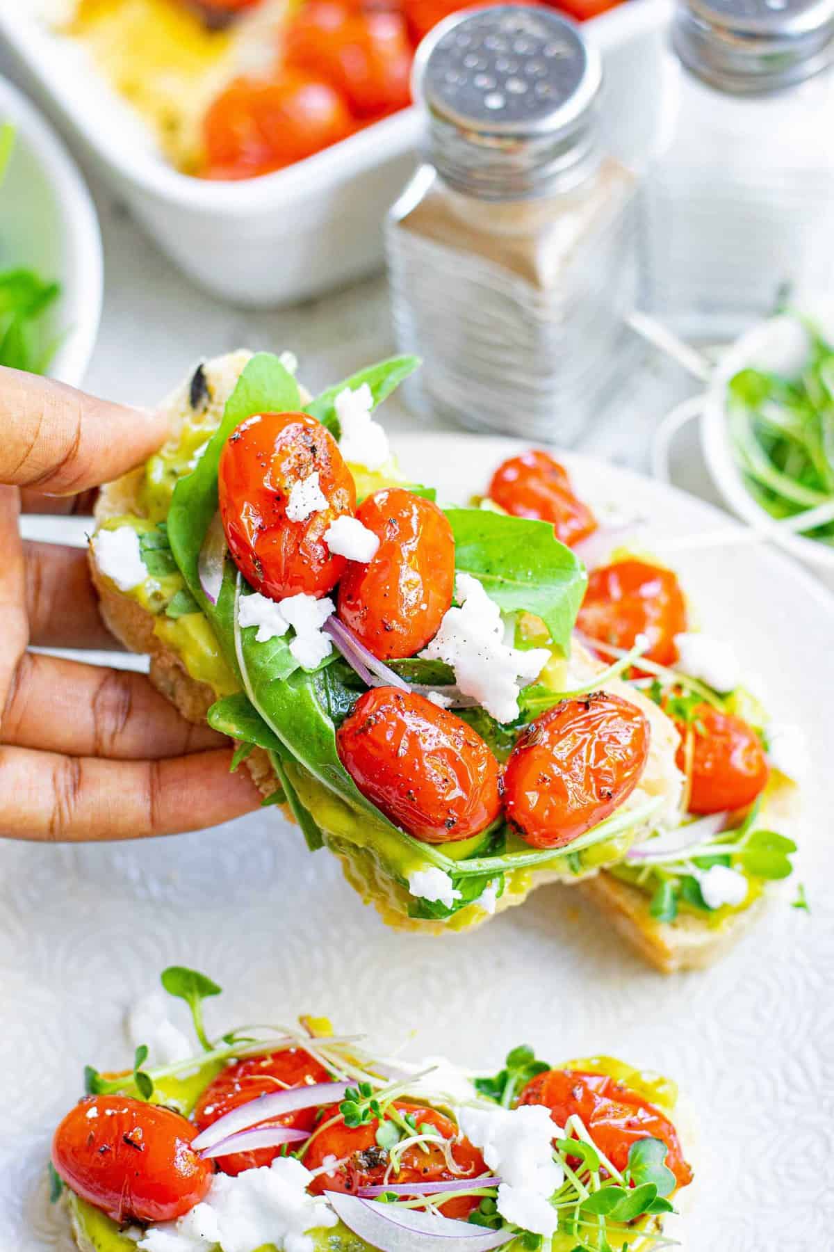 Une main tenant un morceau de pain garni d'avocat, de tomate, de feta et de légumes.