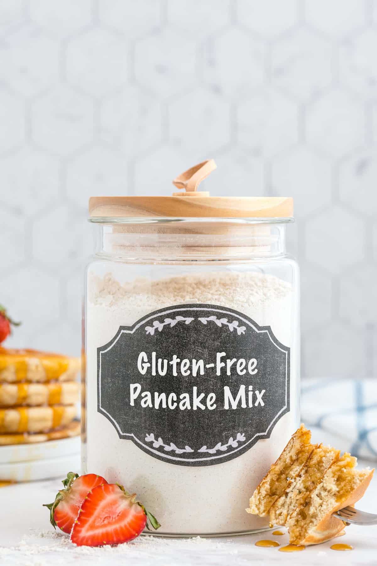 Bocal en verre avec une étiquette noire indiquant 'mélange à pancakes sans gluten'. Pancakes découpés au premier plan.