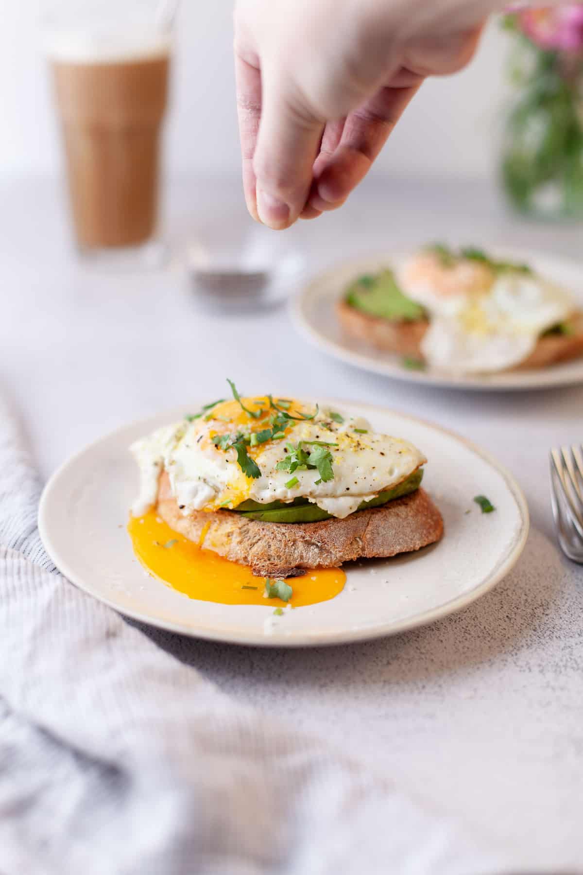 Une main saupoudrant des herbes sur une toast à l’avocat avec œuf.