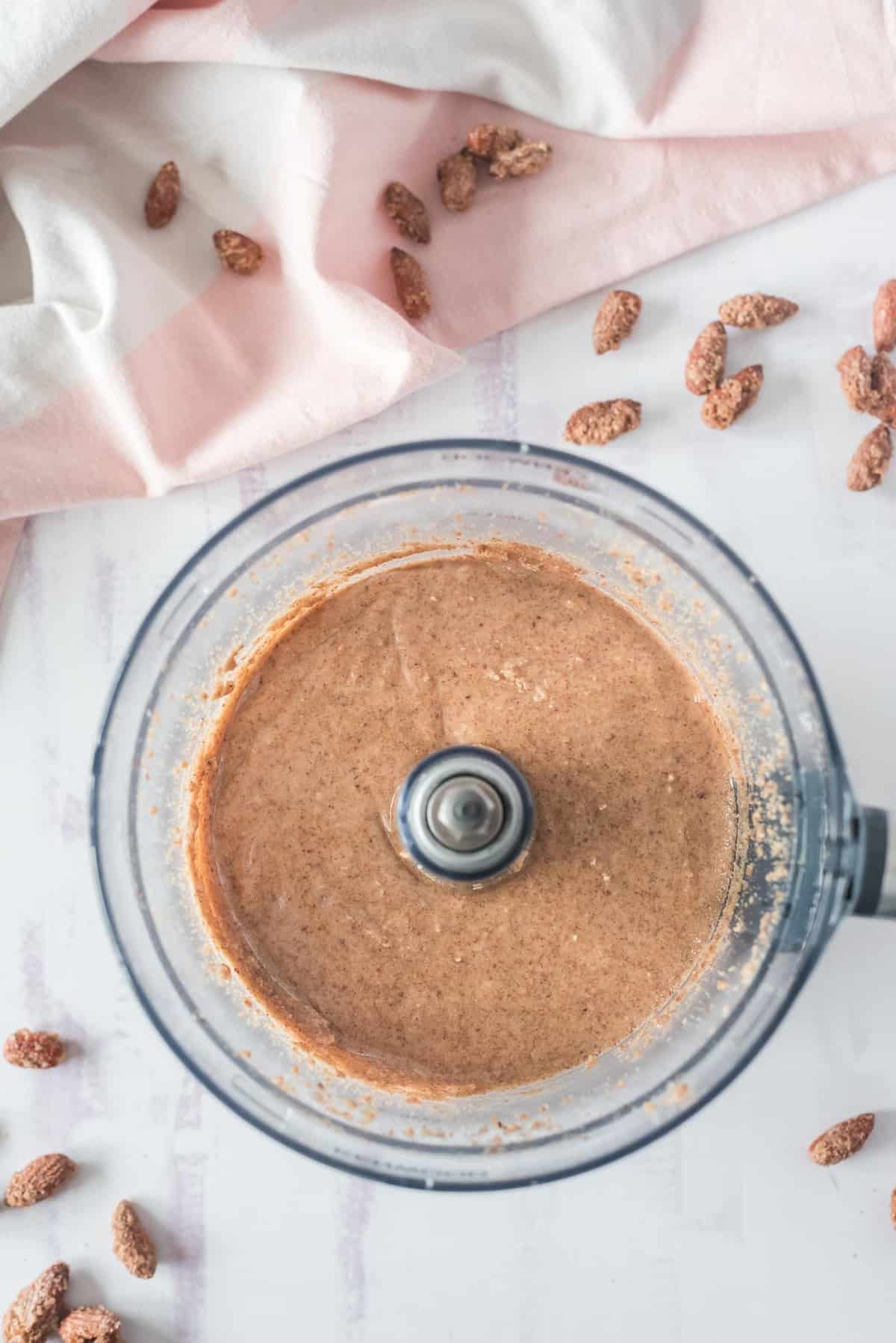 Beurre d'amande crémeux dans un robot culinaire, avec des amandes éparpillées à proximité.