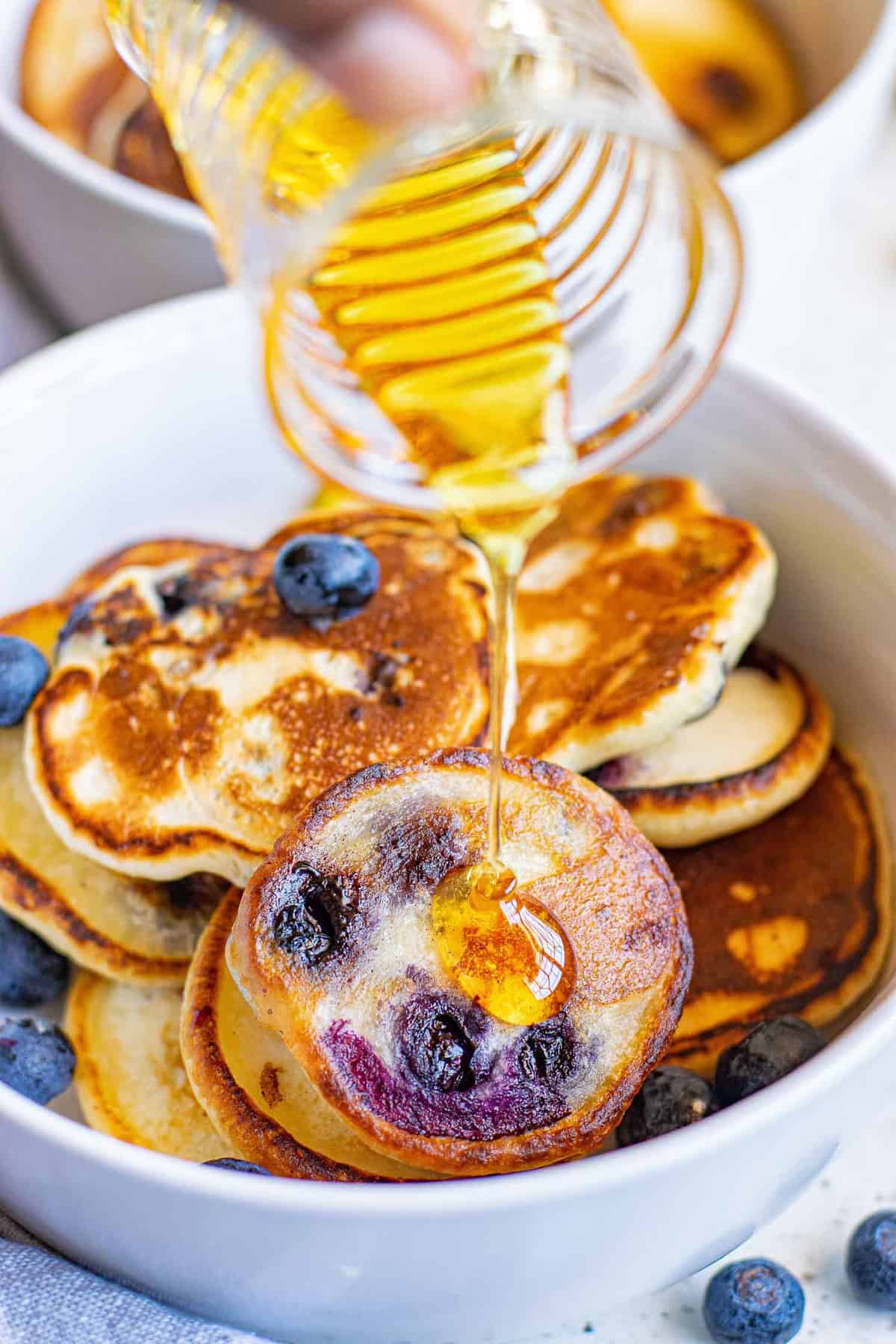 Du sirop versé sur un bol de mini pancakes aux myrtilles.