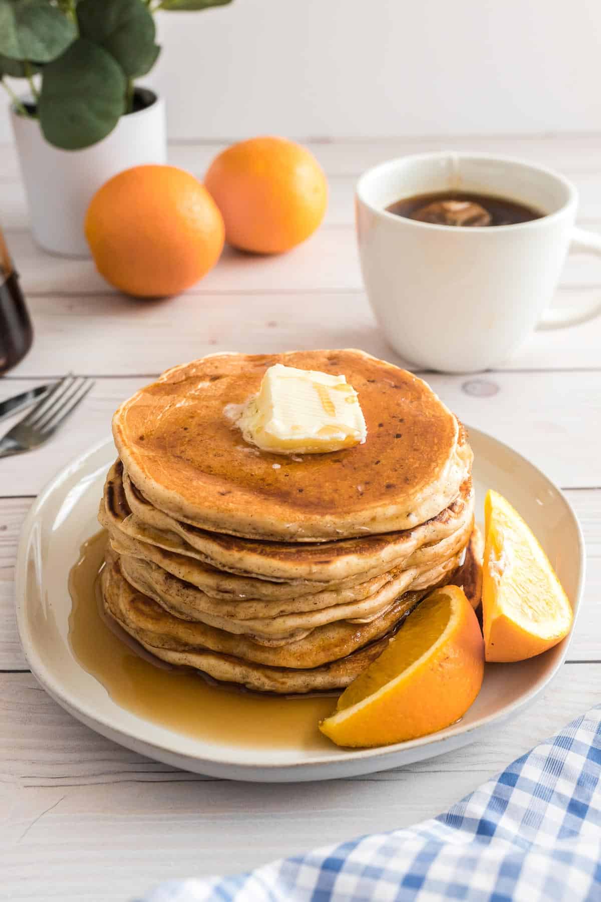 Pancakes et sirop sur une assiette avec des quartiers d'orange, café et oranges en arrière-plan.
