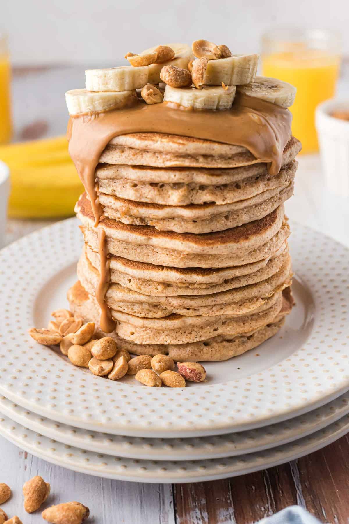 Une très grande pile de pancakes garnie de beurre de cacahuète fondu, de tranches de banane, et de cacahuètes.