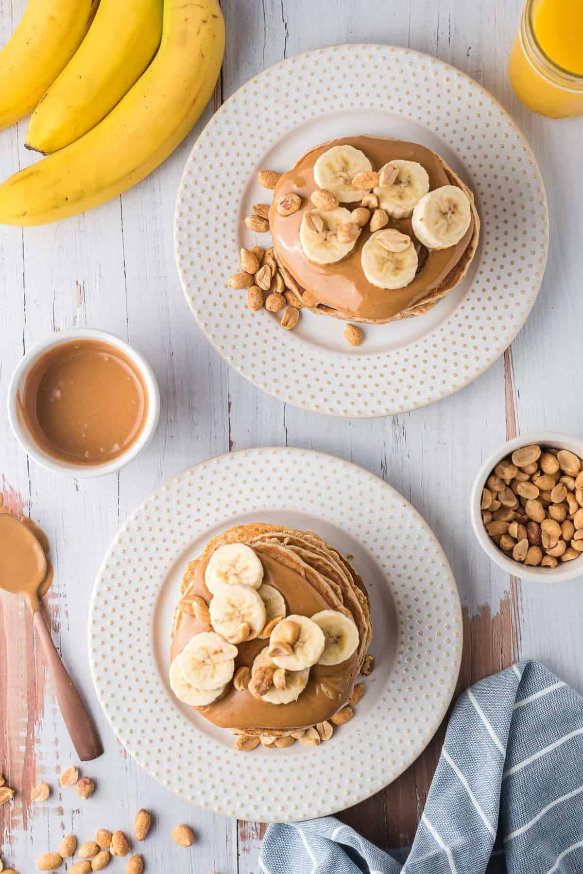 Vue de dessus de deux piles de pancakes sur des assiettes blanches, garnis de beurre de cacahuète, de cacahuètes, et de tranches de banane.