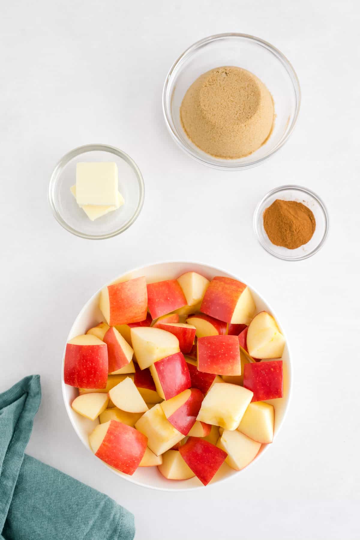 Quatre bols remplis d'ingrédients : pommes en dés, beurre, sucre brun et cannelle.