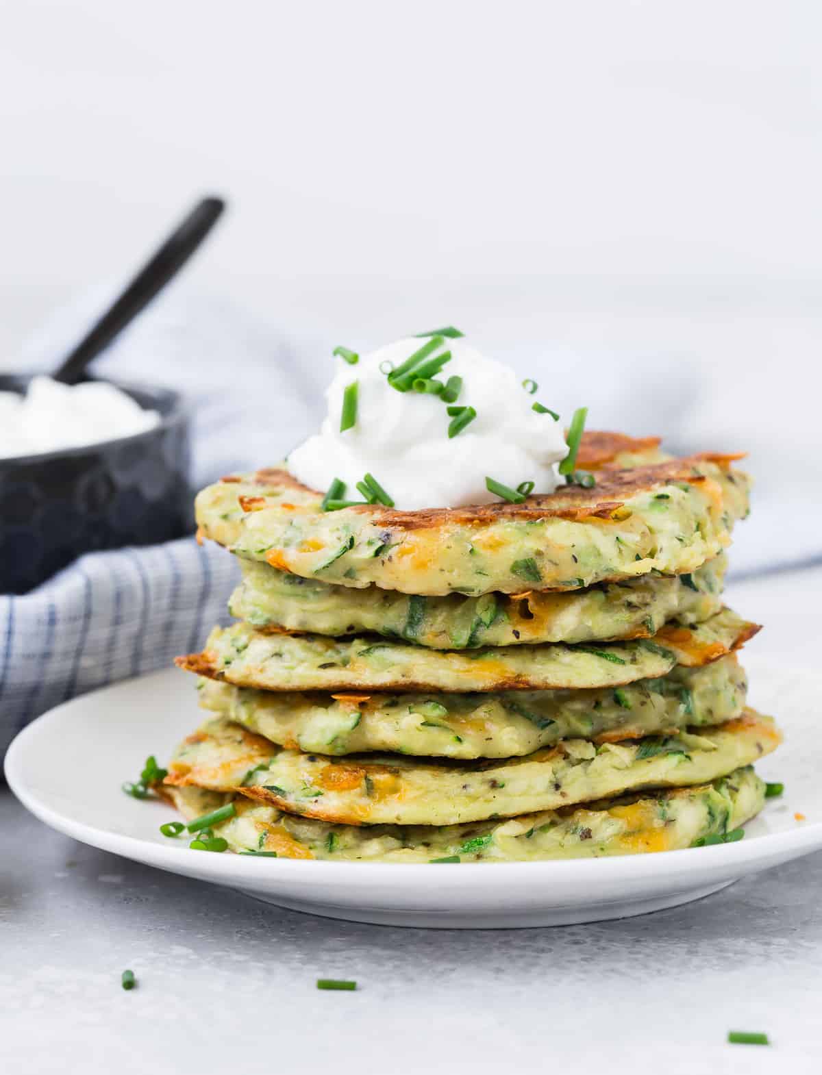 Une pile de pancakes de courgettes sur une assiette blanche, garnie de crème fraîche et de ciboulette.