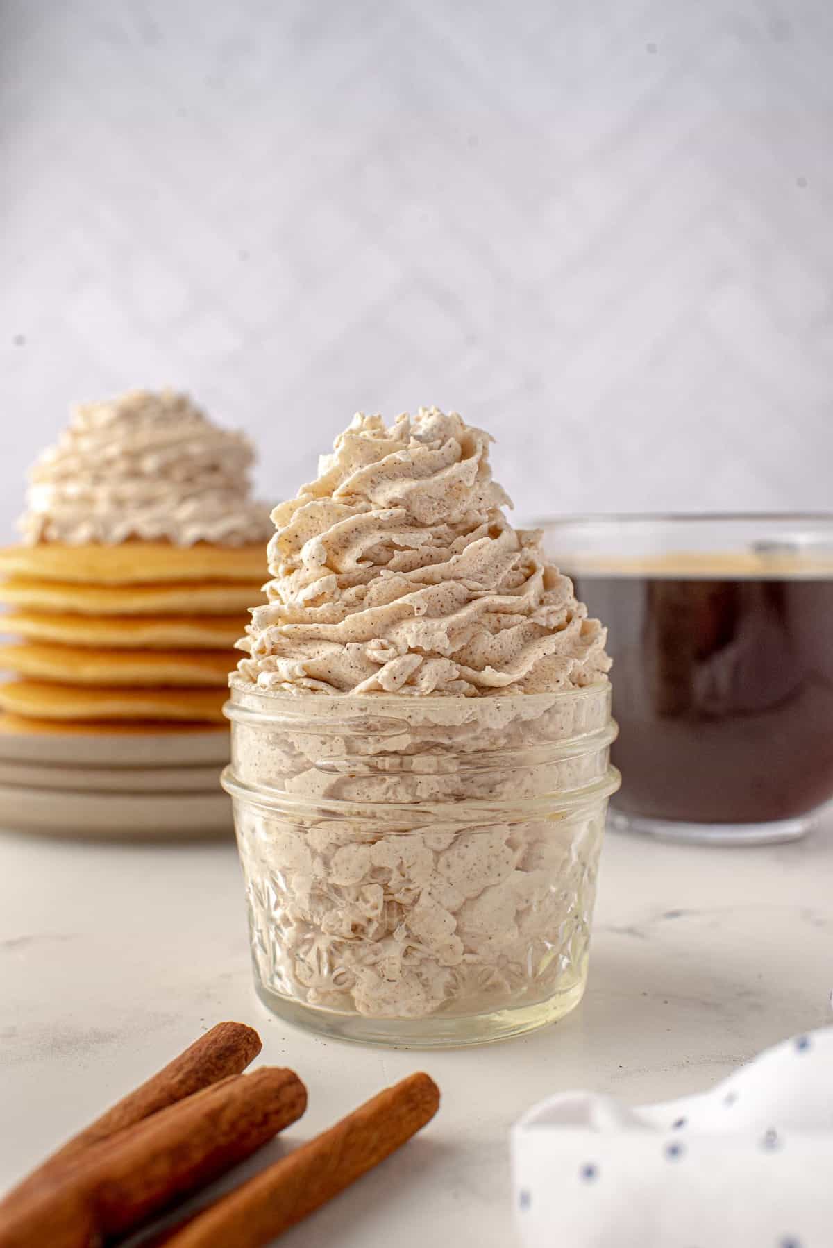 Crème fouettée à la cannelle pipée dans un pot, bâtons de cannelle au premier plan, pancakes en arrière-plan.