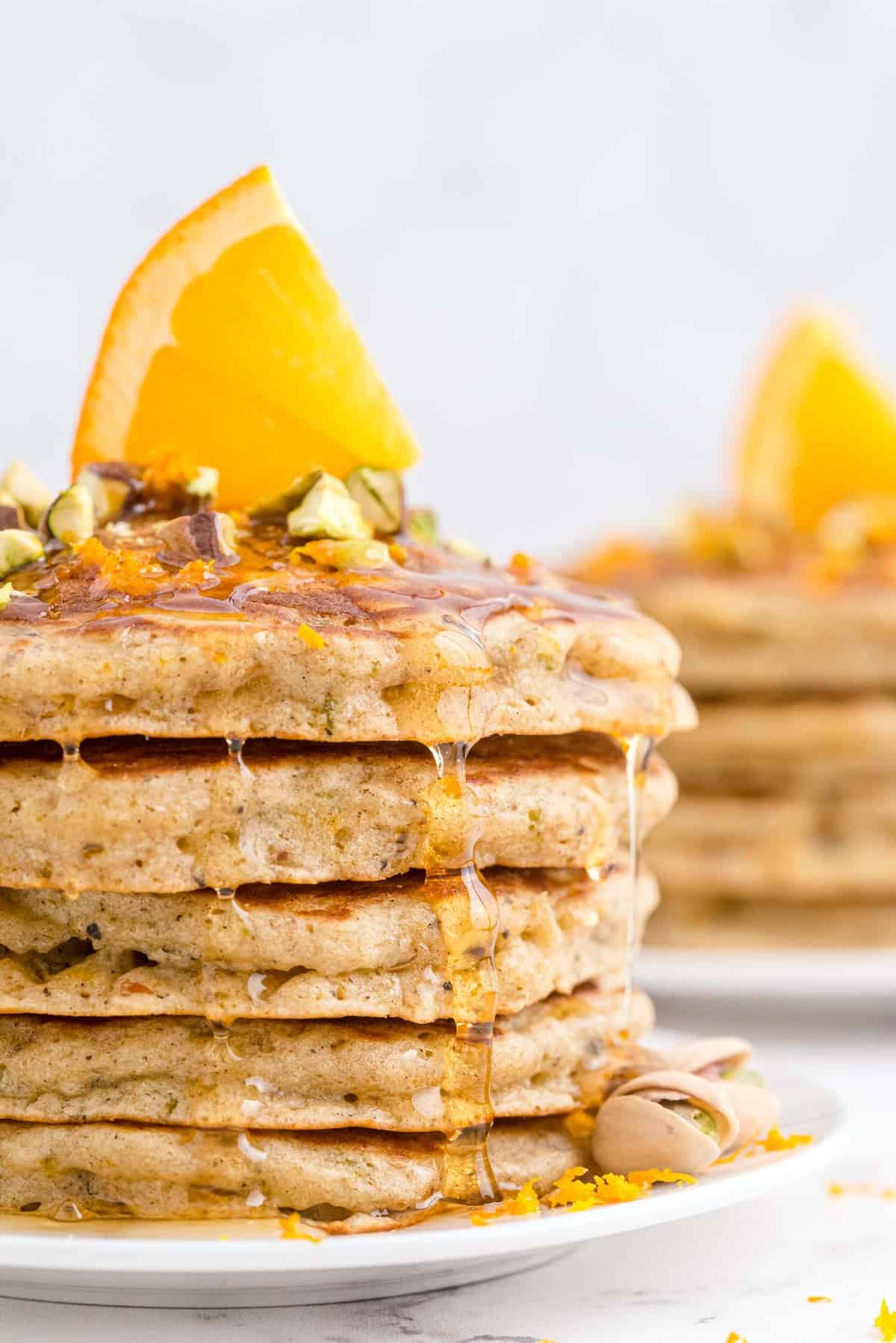 Une pile de pancakes sur une assiette blanche, garnie de pistaches, de miel et d'une tranche d'orange.